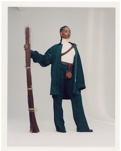 Mereba posing with a broom while wearing an emerald green Jil Sander's oversized shirt, a white knit...