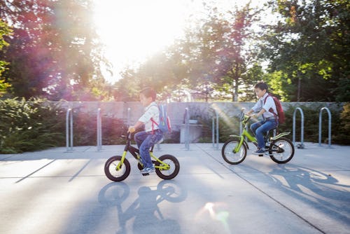 Two little boys with their backpacks riding bicycles on a parking lot next to a Kindergarten