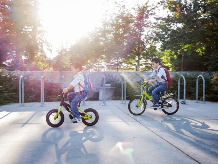 Two little boys with their backpacks riding bicycles on a parking lot next to a Kindergarten