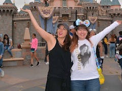 Two sisters wearing Mickey Mouse ears stand in front of Sleeping Beauty's Castle at Disneyland with ...