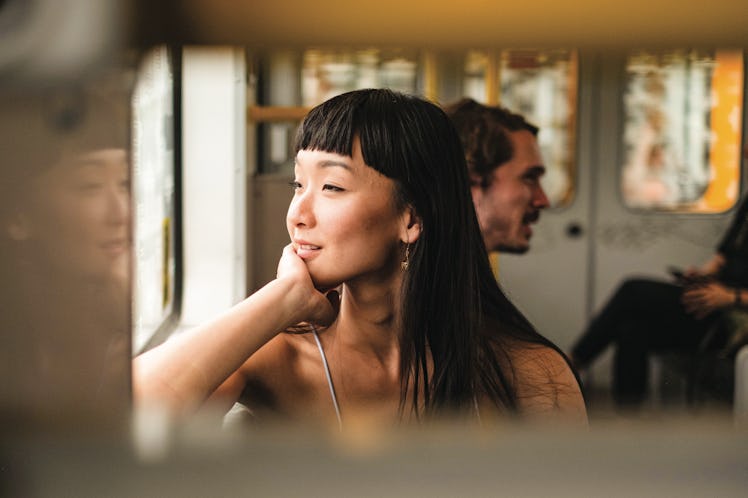 An Asian woman looks out the window of a train while listening to music and on a Contiki trip.