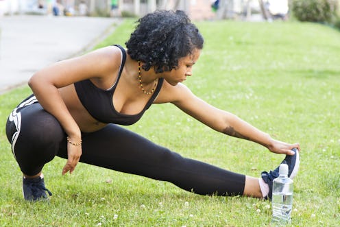 A person wearing a black bra, running leggings, and a rainbow necklace stretches on a grassy field. ...