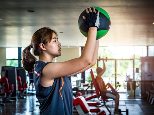 A person wearing a tank top and exercise gloves holds a medicine ball above her head, poised to slam...