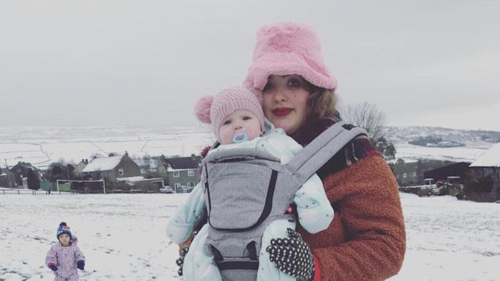 A mother stands in snowy field with a baby in front carrier and toddler in beanie on the ground.
