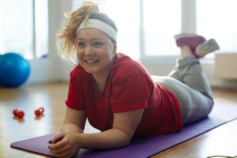 A person in a red t-shirt and white sweatband lays on her purple yoga mat, looking up and smiling. W...