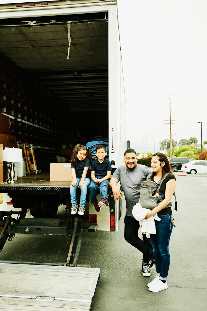A family, including a baby-wearing mother, stand beside a moving truck
