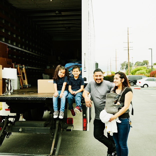 A family, including a baby-wearing mother, stand beside a moving truck