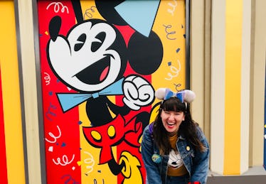 A woman in Mickey ears laughs in front of a Mickey birthday wall at Disneyland.