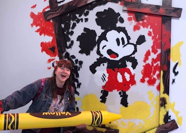 A woman holds up a yellow crayon to a wall with a Mickey Mouse portrait. 