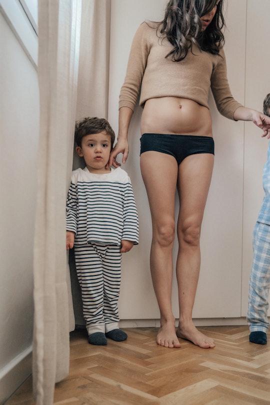 A mother stands with toddler in underpants and sweater