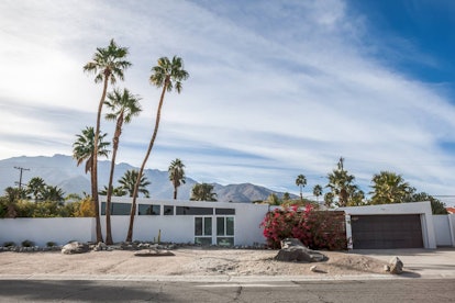 A mid-century modern home in Palm Springs has palm trees in the front yard and mountains behind it.