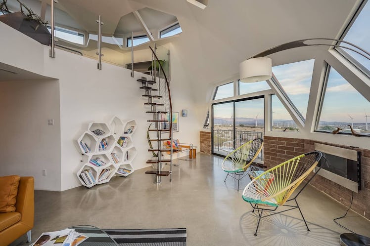 A living room has circular chairs, a small spiral staircase, and an artsy bookshelf in a dome home i...