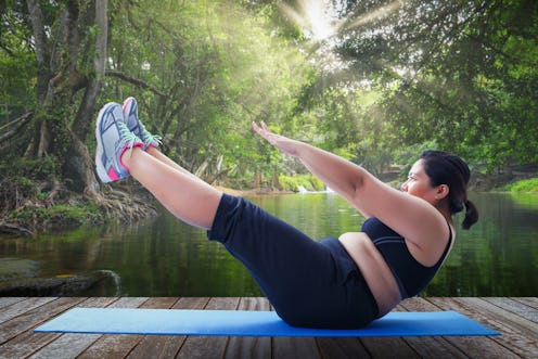 A person holds a core-strengthening pose on a mat in front of a lake. Body-positive Pilates videos c...