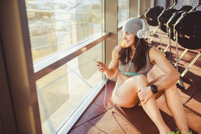 A person wearing a beanie and workout gear looks out the window of her gym while smiling and listeni...