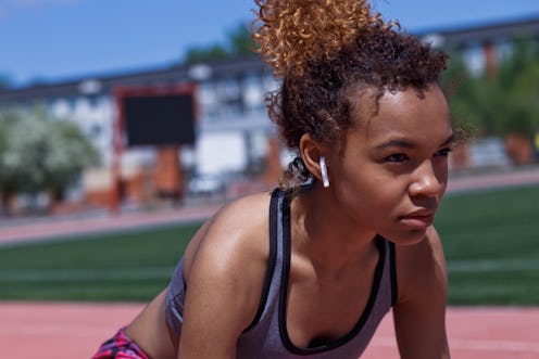 A young Black woman wearing a sports bra and shorts listens to music while preparing to sprint. List...