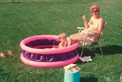 Mother sits with legs in paddle pool, where child plays