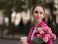 Emily from 'Emily in Paris' wears a hot pink pea coat and holds a coffee cup and bouquet of roses.