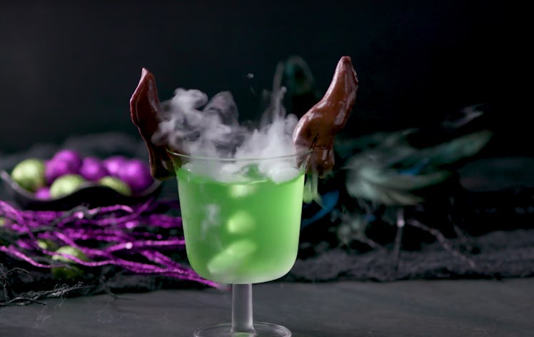 A Maleficent green drink with chocolate horns sits on a table decorated for Halloween.