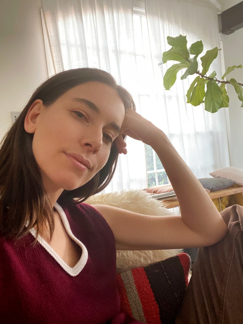 Danielle Haim sitting in her home, in a maroon tank top, in front of a window