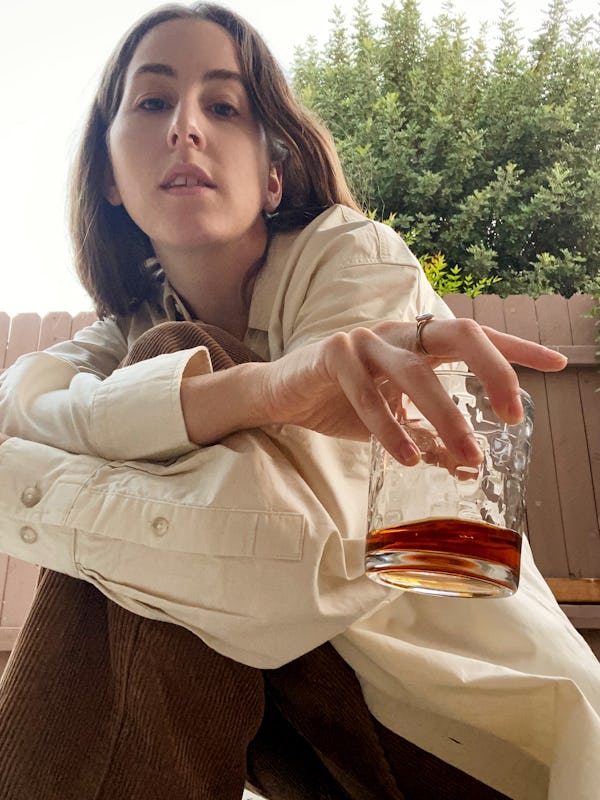 Alana Haim sitting on her balcony in a beige shirt holding a glass of whiskey