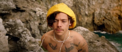 Harry Styles stands on some rocks in the sea with a yellow fisherman's hat on. 