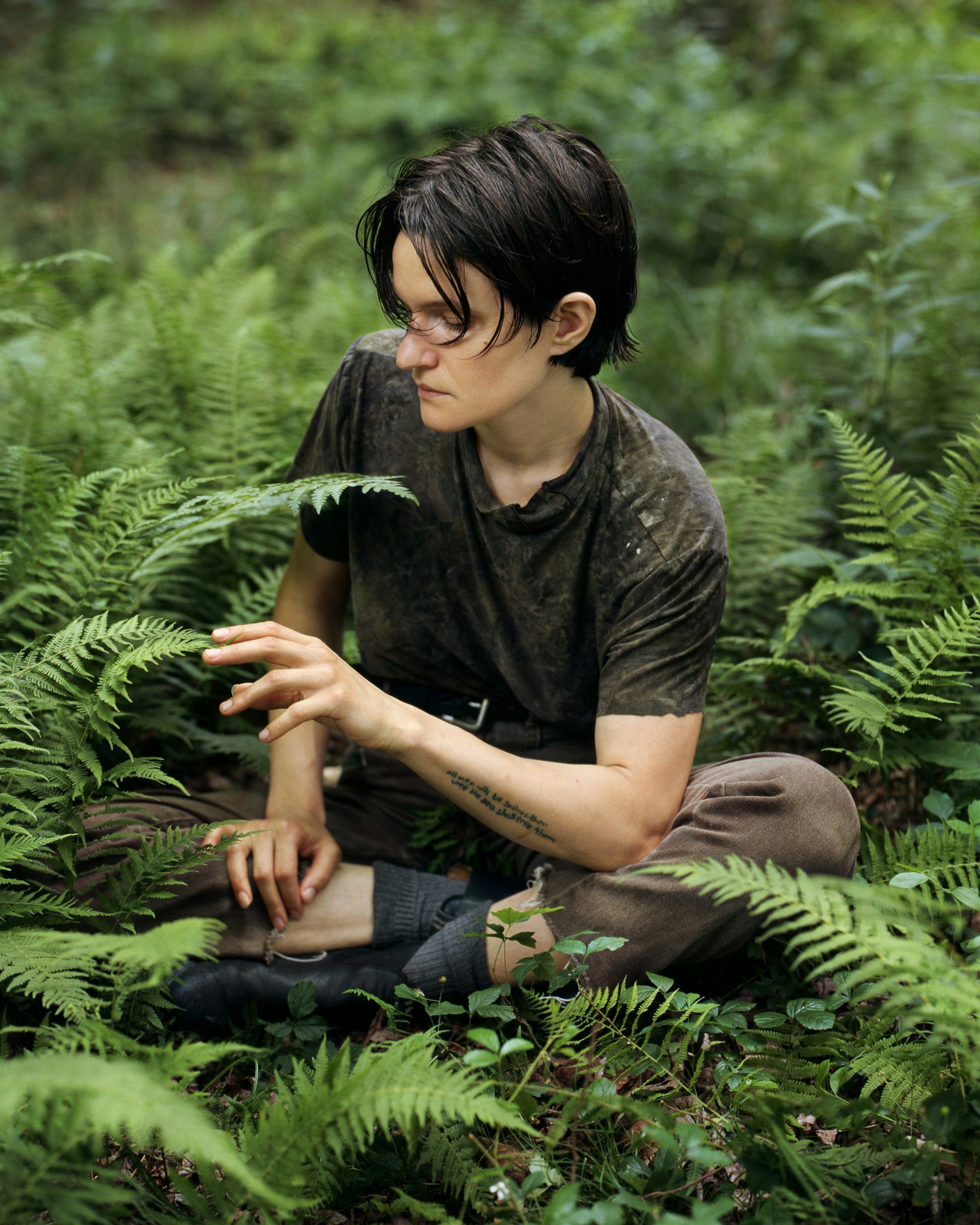 Singer-songwriter Adrianne Lenker sits among fern on the forest floor, gently touching the tip of a ...