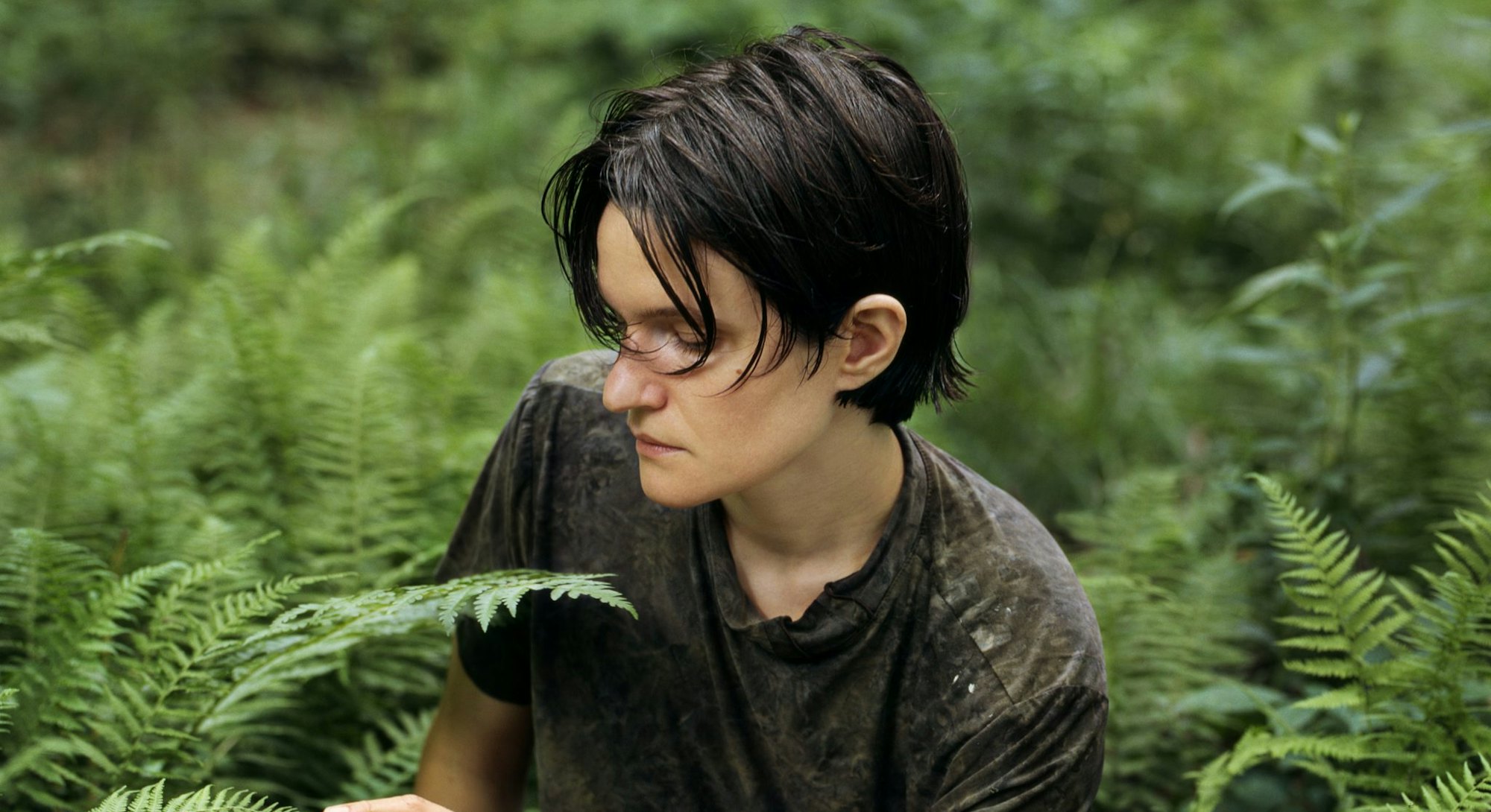 Singer-songwriter Adrianne Lenker sits among fern on the forest floor, gently touching the tip of a ...