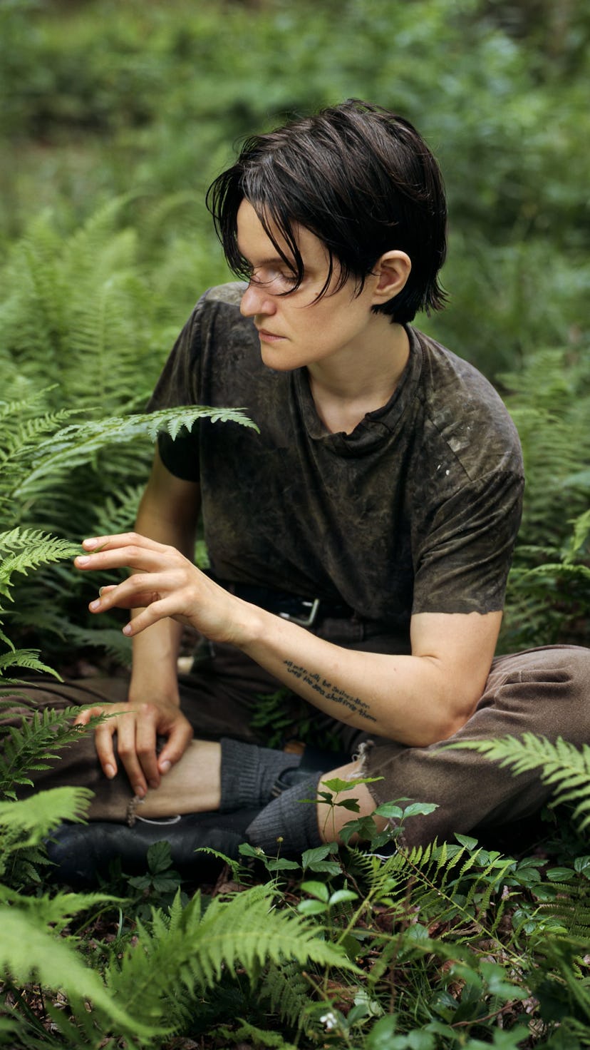 Singer-songwriter Adrianne Lenker sits among fern on the forest floor, gently touching the tip of a ...