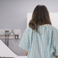 A woman sits on a doctor's examining table in a hospital gown. The impact of delayed health care dur...