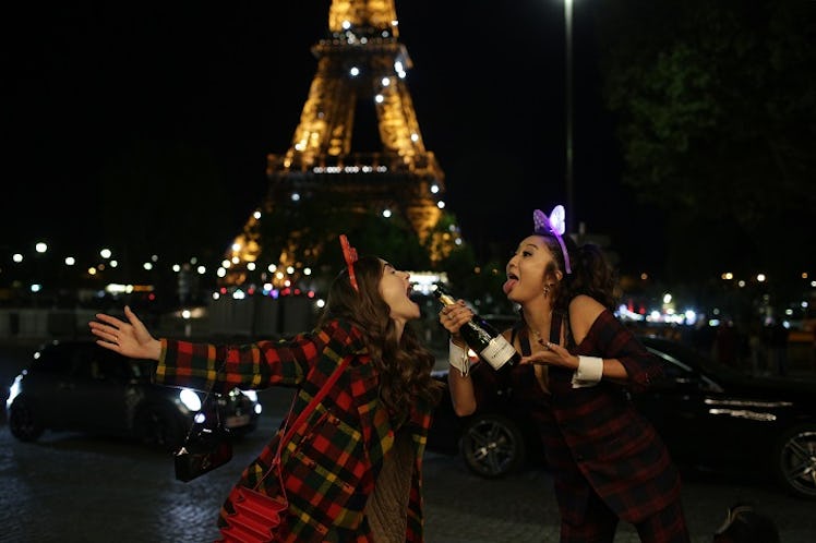 Emily and Mindy from 'Emily in Paris' wear plaid and make silly faces while holding a champagne bott...