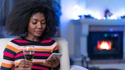 A woman with an afro scrolling on her phone with a glass of wine, as she is alone during the pandemi...