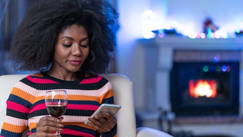 A woman with an afro scrolling on her phone with a glass of wine, as she is alone during the pandemi...