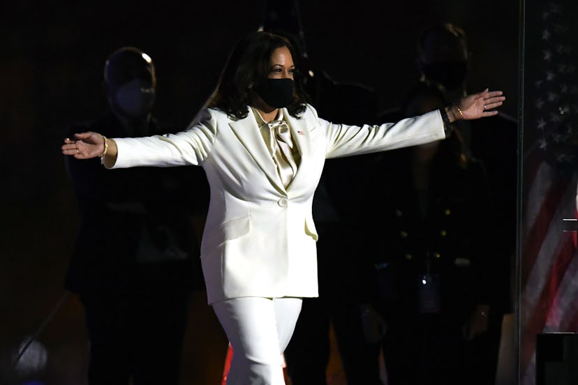 Vice President-elect Kamala Harris wears a white suit, a black mask, and her arms are spread out wid...