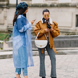 Two women stand outside, one is in a blue outfit the other in jeans and a suede jacket