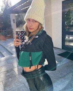 Hailey Bieber holds a cup of coffee while standing outside in the cold in a white hat, a short jacke...