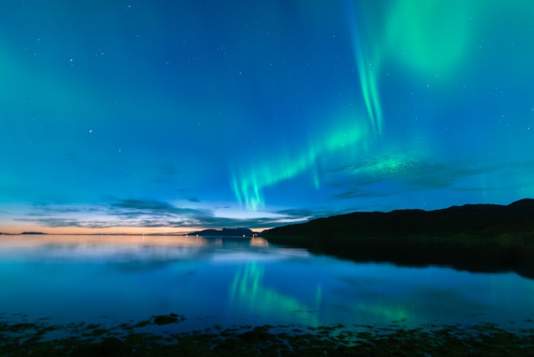 The Northern Lights light up a blue sky over a lake in Norway.