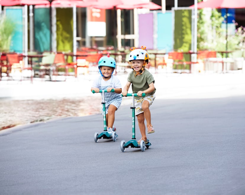 Two kids in helmets on Micro Kickboard scooters, riding down the street