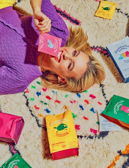 Emma Chamberlain lays amongst the Chamberlain Coffee products on a colorful rug.