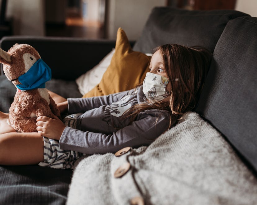A little girl sits on her couch at home with a stuffed bunny; they are both wearing face masks.