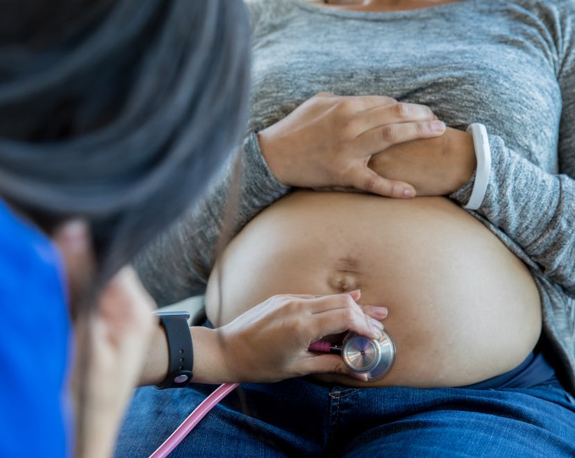 Close-up of a doctor's hands holding a stethoscope to a bare pregnant belly.