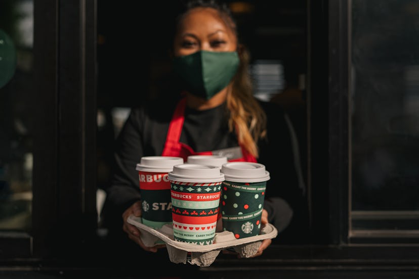 A woman with brown hair and eyes, wearing a green face mask, holding four Starbucks cups in a tray, ...