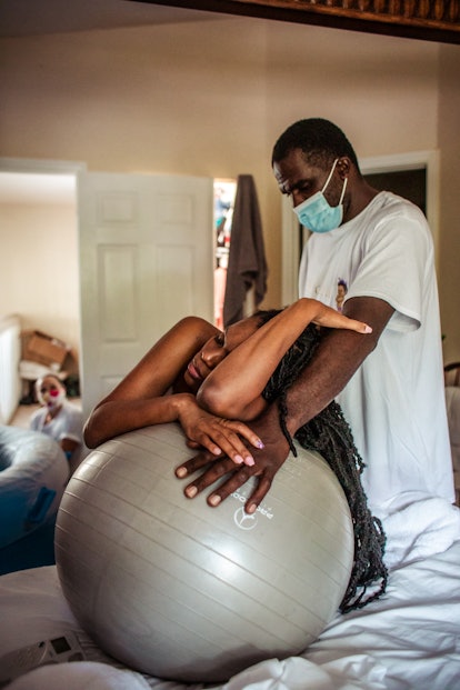 a woman labors with a birthing ball while her husband supports her as he wears a mask