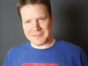 A white man with dark short hair wearing a T-shirt with a logo on his chest, which is the written AS...