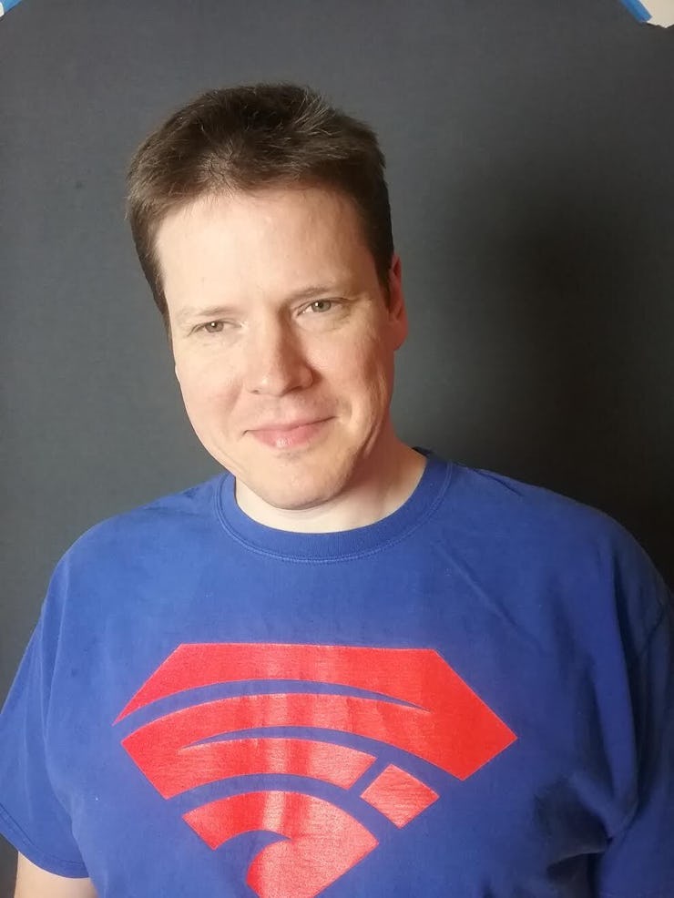 A white man with dark short hair wearing a T-shirt with a logo on his chest, which is the written AS...