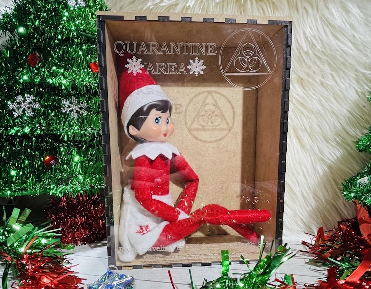 An Elf on the Shelf sits in a quarantine Christmas box from Etsy, surrounded by tinsel Christmas tre...