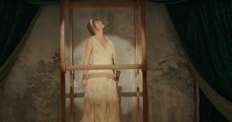 Taylor Swift stands in a glass box while wearing a white dress and holding a ukulele in the "willow"...
