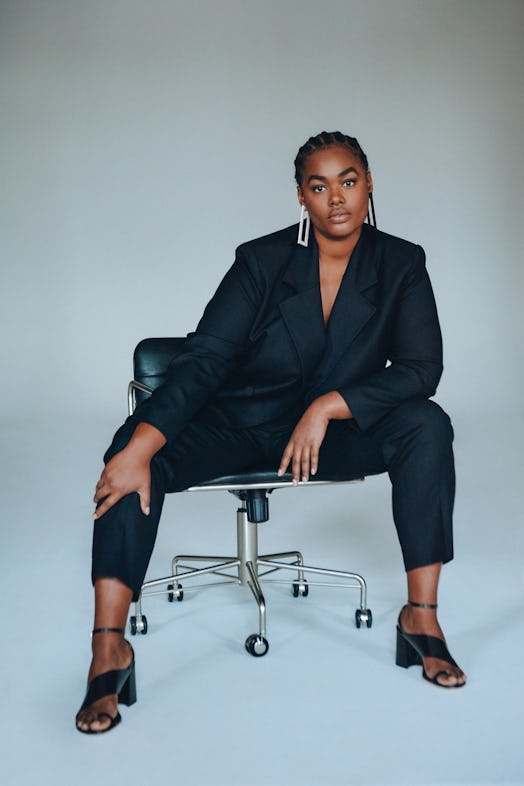 A plus-size model sitting and posing in a black blazer and in black pants