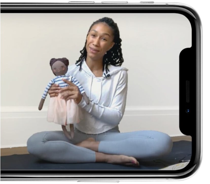 A phone screen shows a woman with braids sitting in a yoga post, holding up a Brown baby doll to the...