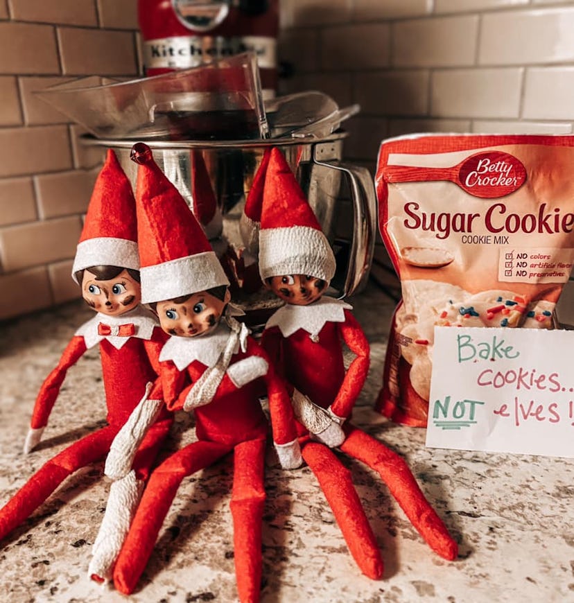Three Elf on the Shelf dolls sit on a kitchen counter, variously bandaged, with a sign that reads "B...