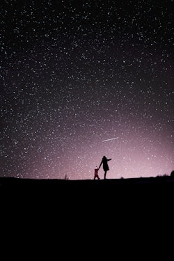 A mother and child in a dark field, seen from afar, under a glorious night sky filled with stars.